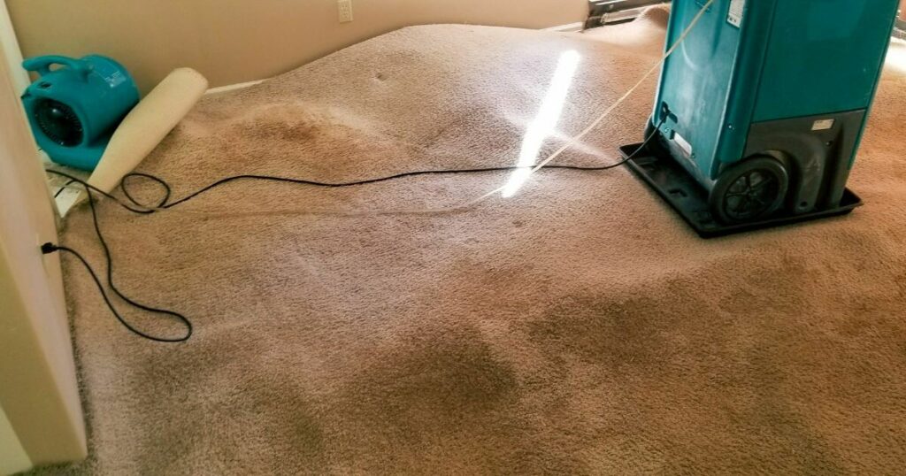 A room undergoing water damage restoration with professional equipment, including a dryer and dehumidifier, set up to dry the wet carpet.